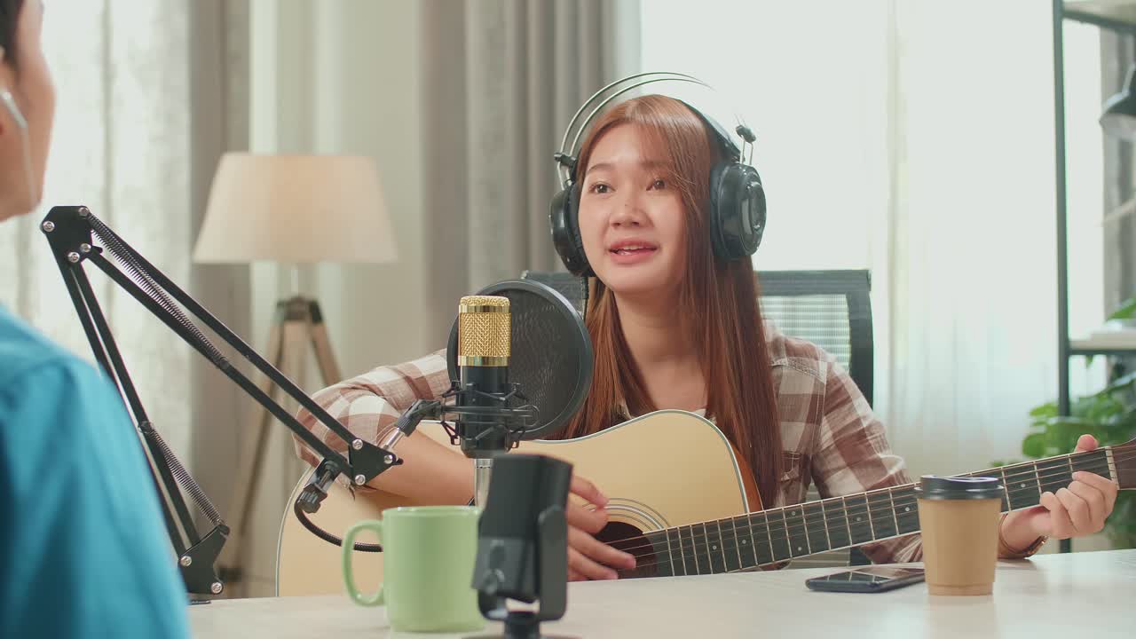 Over Shoulder View, Young Asian Woman In Headphones Playing The Guitar And Singing Into Microphone While Recording Podcast With Male Host In Studio