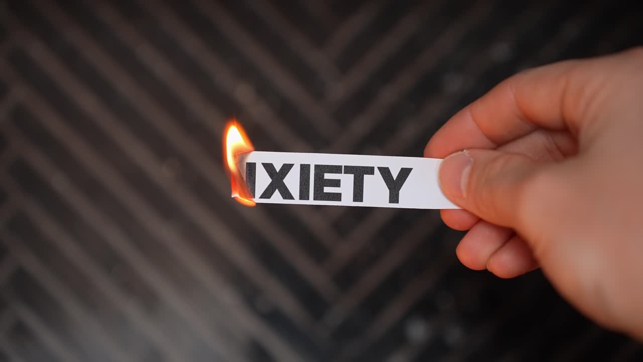 Close-up hand burns a paper label reading “ANXIETY,” symbolizing stress, panic, and mental health struggle, with dark background and copy space for therapy or recovery themes