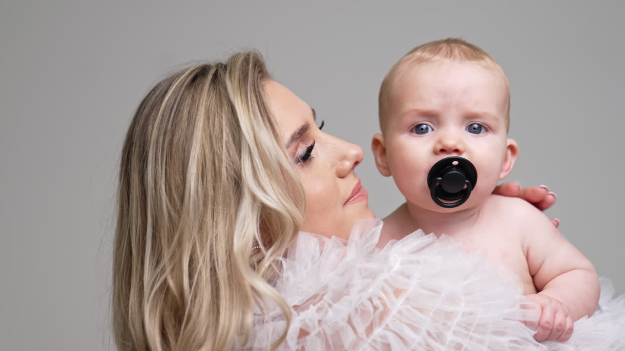 Blonde woman stroking her tiny son by the head. Mother kisses her child sucking a pacifier. Lose up. White backdrop.