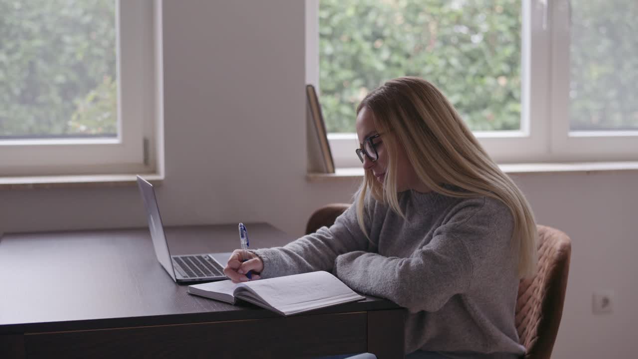 En ung kvinne med briller studerer ved et skrivebord med laptopen sin og tar notater i en notatbok. Naturlig lys fra vinduet skaper en rolig og fokusert hjemmestudiestemning.