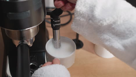 woman uses cappucinator making milky foam. lady whips milk in pitcher with hot steam pipe from coffee machine at table. process of making cappuccino