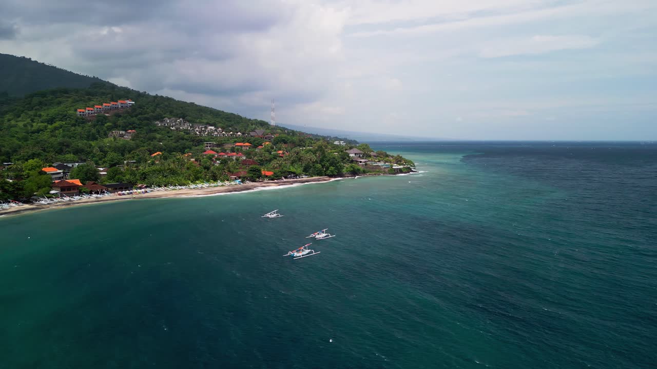 Filmato aereo con drone di barche da pesca tradizionali ancorate sulla riva di una laguna blu, accanto a una costa verdeggiante collinare dell'Indonesia con acqua turchese e uno scenario di spiaggia tropicale con onde