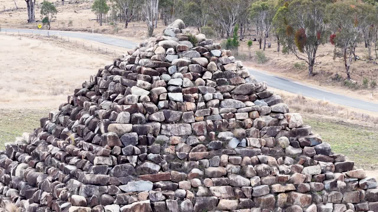 Drone camera steadily approaches a large granite rock pyramid in a dry, open landscape with scattered trees and overcast daylight