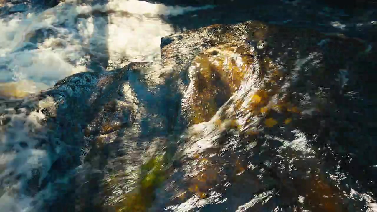 La luz del sol atrapa el agua que fluye mientras se desliza sobre una roca lisa, creando un vívido juego de brillo, textura y movimiento en un detalle abstracto cercano de un arroyo.