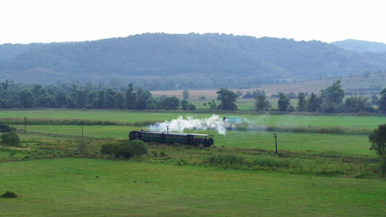 A historic steam train with passenger cars makes a scenic journey through a lush green valley. The locomotive puffs white steam as it travels through the countryside with forested hills