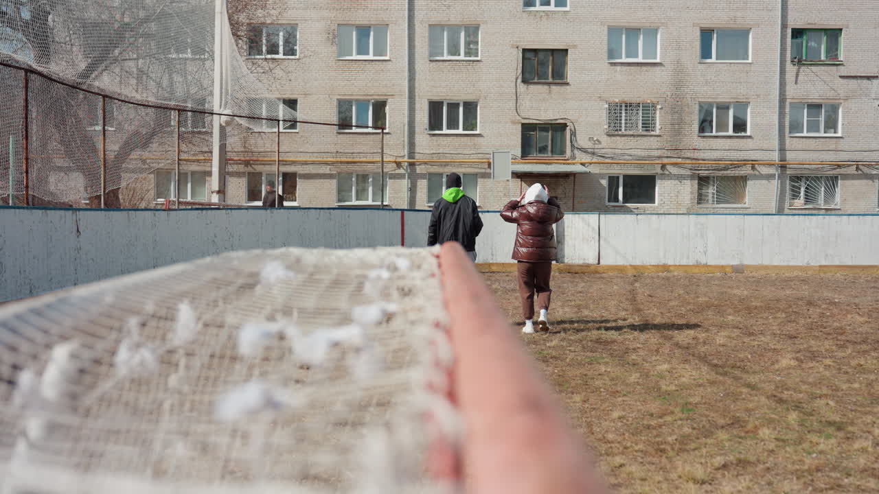 흰색 10대 소녀가 골대 근처에서 공을 줍기 위해 몸을 굽히고 있고, 동료는 등을 돌리고 있으며, 낡은 경기장, 뒤로는 아파트 단지, 집중된 움직임과 작은 승리, 캐주얼한 겨울 의상과 솔직한 운동 모습