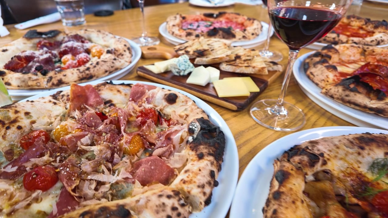 La telecamera si allontana rivelando diverse pizze gourmet, un tagliere di formaggi e vino rosso su un tavolo di legno.
