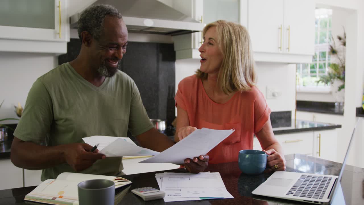 feliz pareja mayor diversa usando computadora portátil y pagando facturas en la cocina