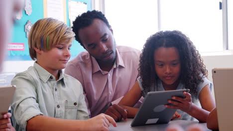 Male-teacher-helping-two-young-kids-using-tablet-in-class