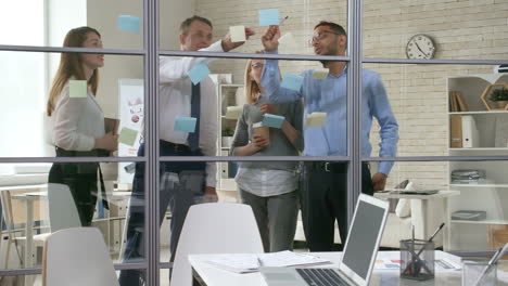 equipo de negocios de mentores senior y colegas jóvenes escribiendo y pegando notas adhesivas en la pared de vidrio en la oficina y hablando de planes 1