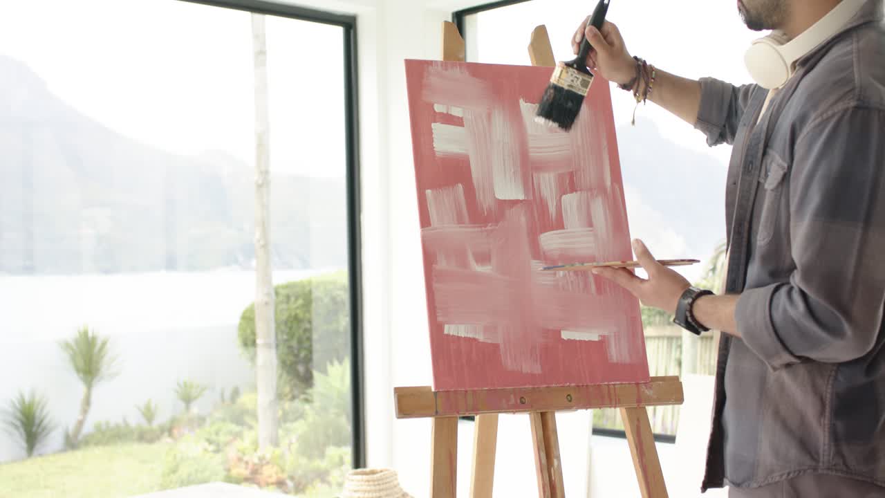 画家が筆を絵の具に浸し、窓辺の赤いキャンバスに白い交差するストロークで格子状の模様を描いている。