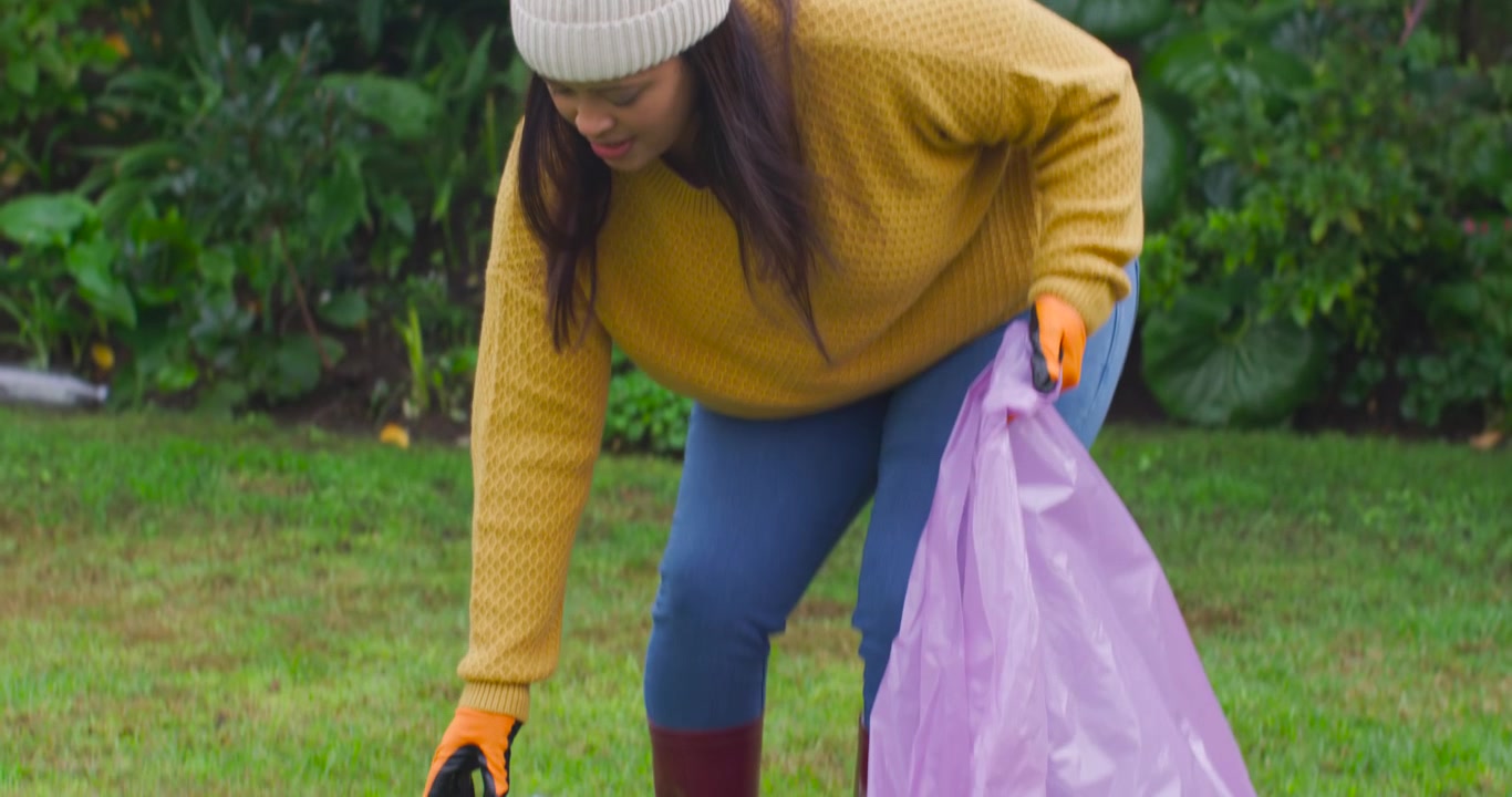 African American women volunteering on lawn during cleanup, picking water bottle into purple bag