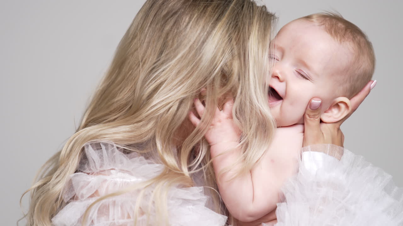 Mom playing with her adorable baby. Blonde woman holding her tiny son and kisses him on shoulders and cheeks. White backdrop.