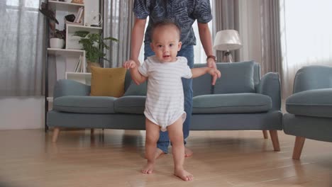 asian father encouraging smiling baby son to take first steps and walk at home