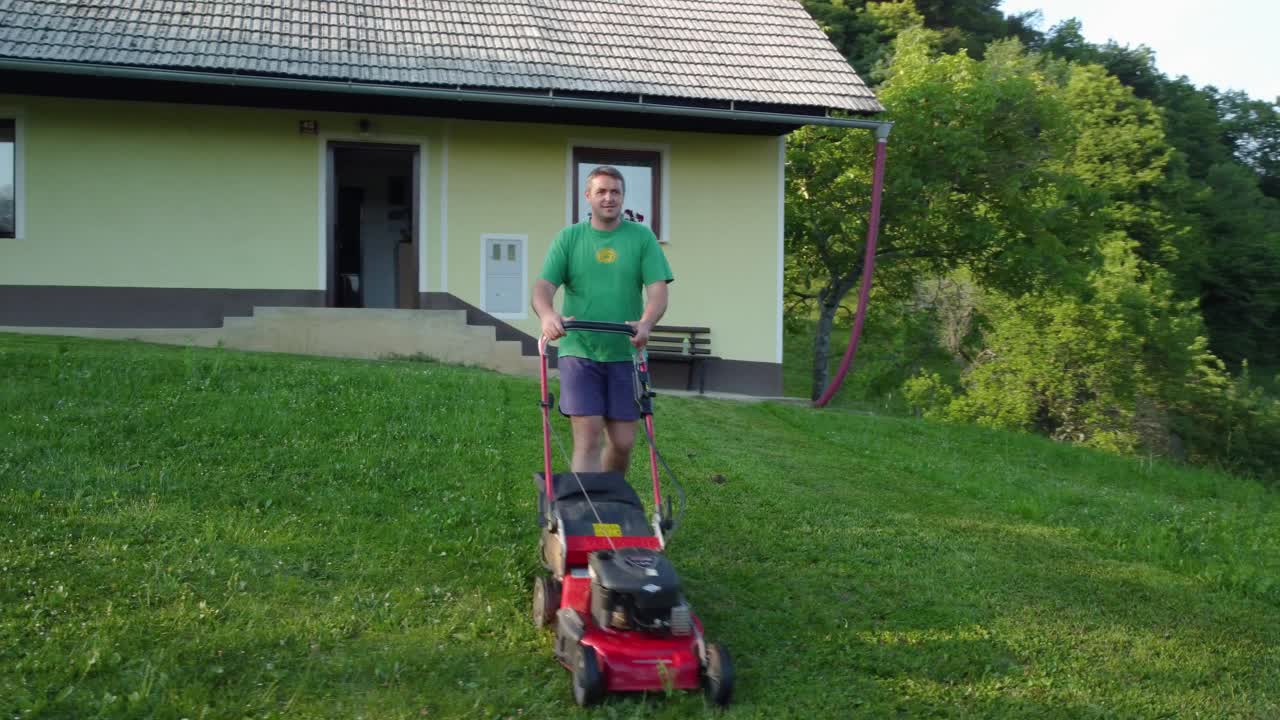 Szczęśliwy mężczyzna w niebieskich szortach i zielonej koszuli kosi zielony trawnik przed swoim domem