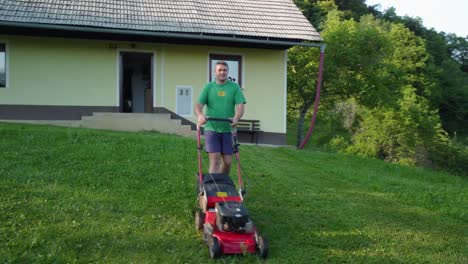 happy man in blue shorts and green shirt mowing green lawn in front of his house