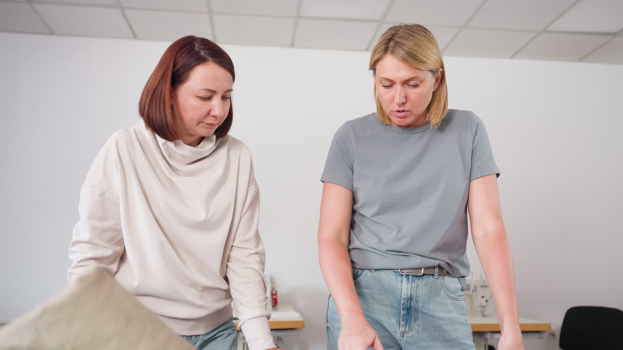 Suit maker explaining fabric details to attentive lady standing beside her, discussing textile selection for fashion project in creative workspace with sewing machines and tools visible in background