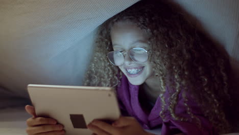 linda chica de raza mixta escondida debajo de una manta, leyendo un libro electrónico