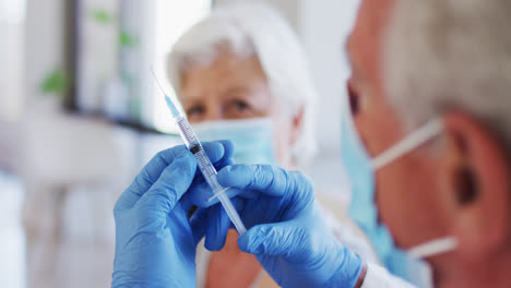 médico caucásico masculino sosteniendo una jeringa mientras una mujer caucásica senior con máscara facial sentada con sl