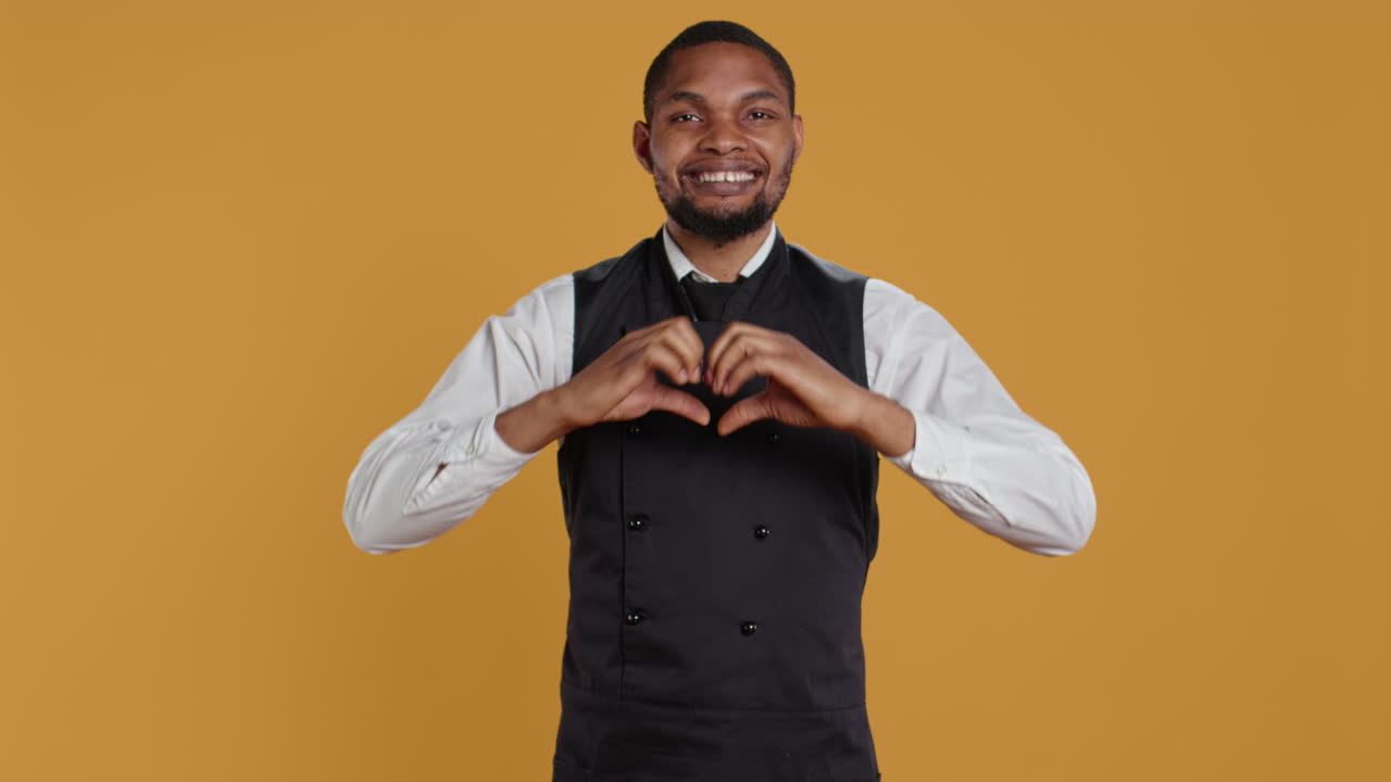 Waiter in apron and uniform showing a heart shape sign in studio