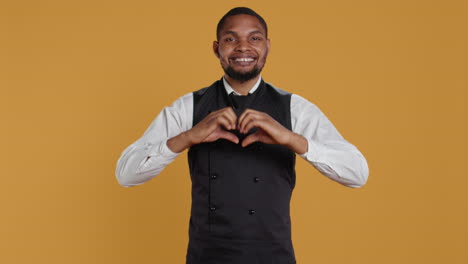 Waiter-in-apron-and-uniform-showing-a-heart-shape-sign-in-studio