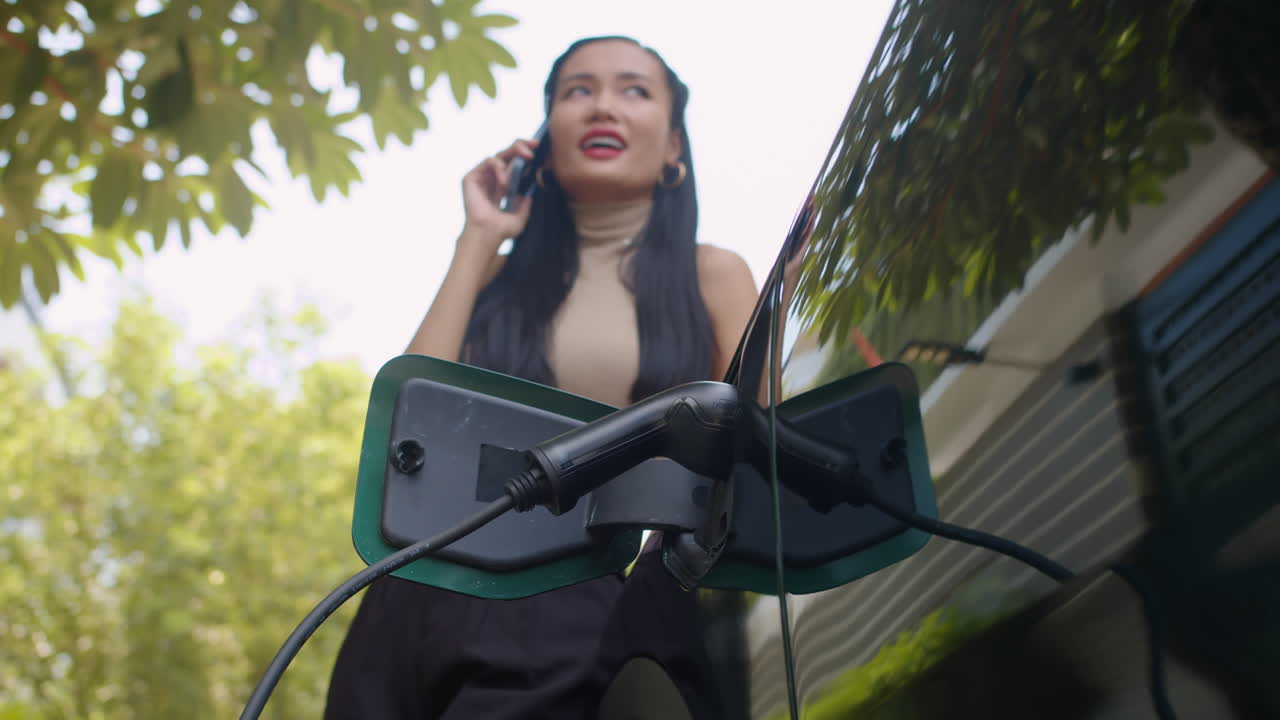 Woman charging electric vehicle while checking smartwatch and talking on phone