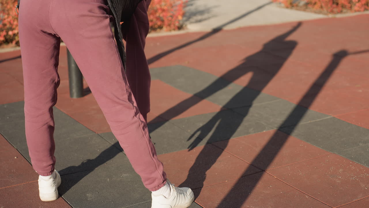 Back View Of Fitness Coach Bending Sideways On Outdoor Gym Court Pavement, Reaching Down To Touch Toes Under Sunlight, Shadow Stretching Across Patterned Tiles, Environment With Red Shrubs