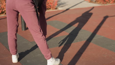 back view of fitness coach bending sideways on outdoor gym court pavement, reaching down to touch toes under sunlight, shadow stretching across patterned tiles, environment with red shrubs