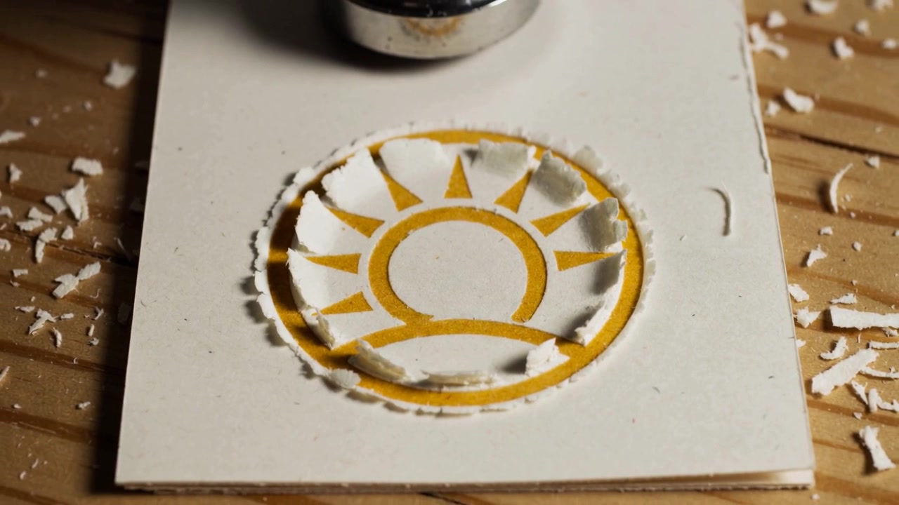 Lowering metal punch cutting center circle from white card on table, exposing yellow sun, chips