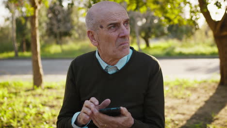 bald elderly man sitting in the park using his smartphone