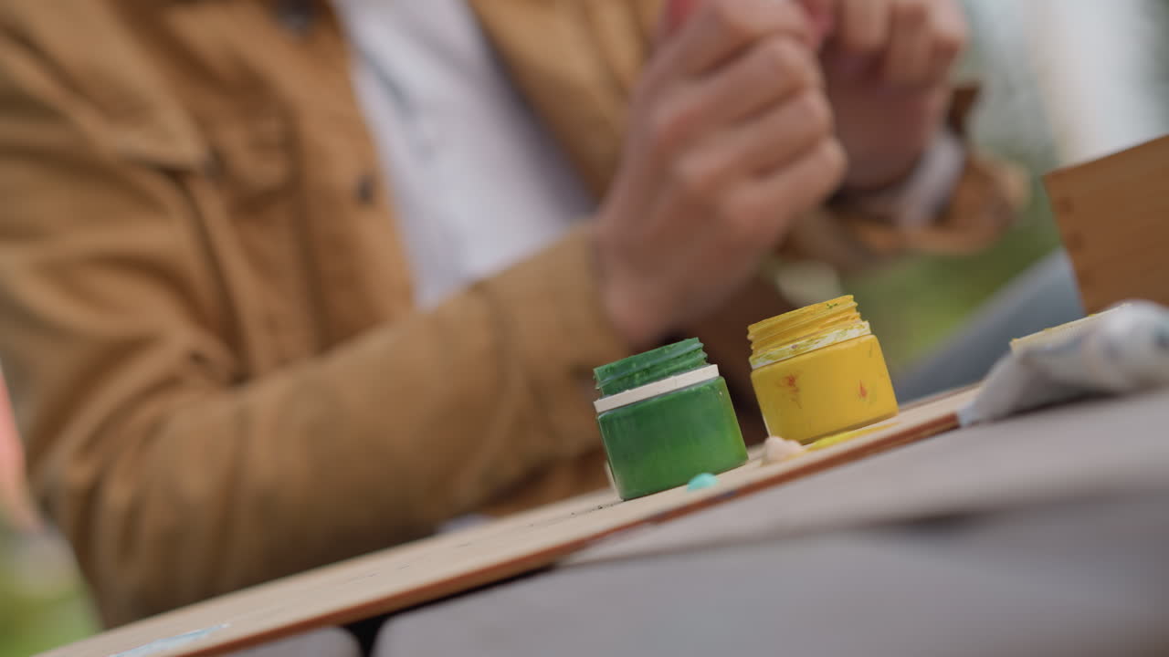 closeup hands of white street artist arranging paint jars on sketchboard, brown jacket, green and yellow acrylic jars, lid placement, palette cloth, outdoor park bokeh, focused tactile process
