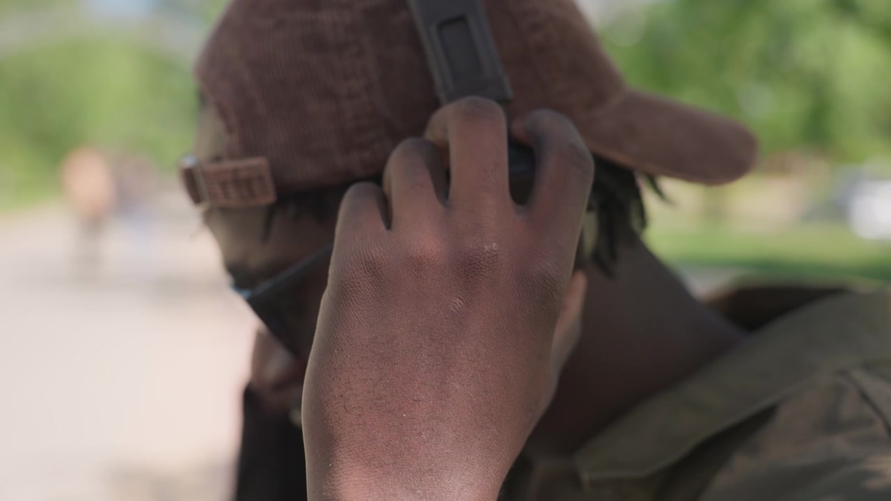 Close up of young man wearing headphones and holding them with one hand focusing on his ear and device. subject is outdoors in daylight surrounded by greenery creating soft natural lighting