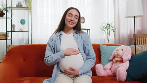 Retrato-De-Una-Futura-Madre-Sonriente-Sentada-En-Un-Sofá-Tocando-Su-Barriga-Embarazada-Esperando-Un-Bebé-En-Casa