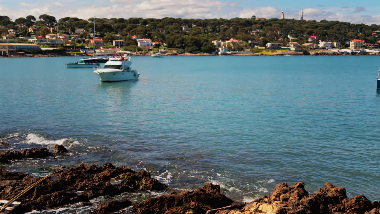 Waves crashing on the rocks on the shore with a boat floating on the sea and the town of Bandol, France on the background