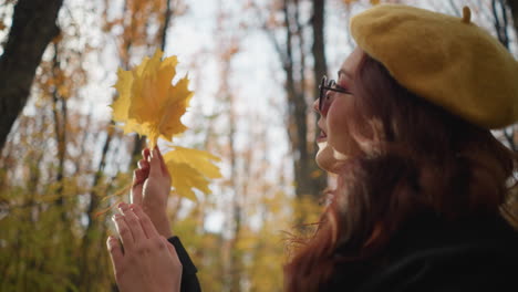 elegant lady in yellow beret and sunglasses twirling vibrant autumn leaves in hand as sunlight reflects on face, surrounded by tall trees in a serene forest filled with golden fall foliage