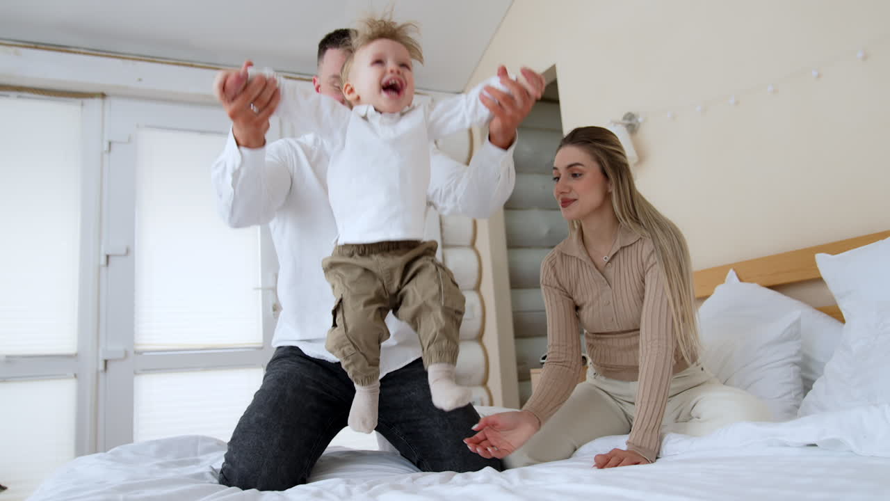 Dad lifts his baby boy jumping happily on the bed. Mother sitting beside touching her son tenderly.