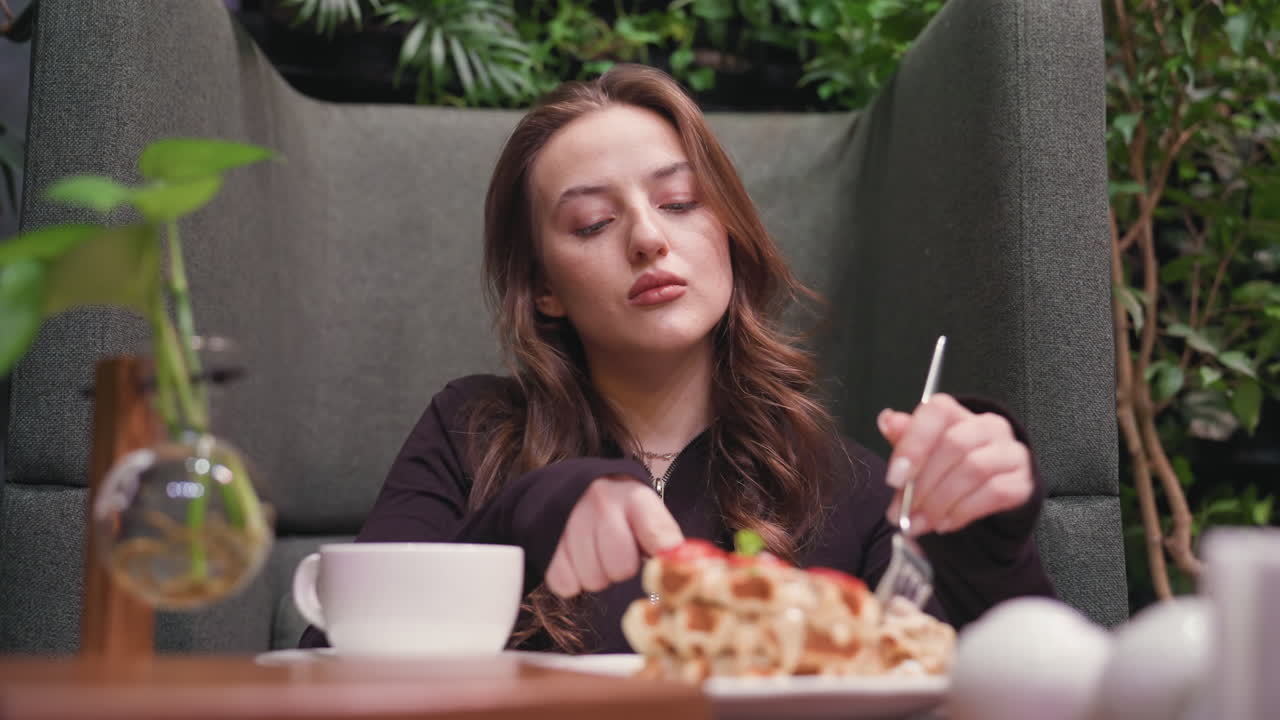 Lady gently cuts waffles into small pieces using knife and fork while seated at cozy cafeteria table with hot drink, decorative plant, and lush greenery in background enhancing natural ambiance