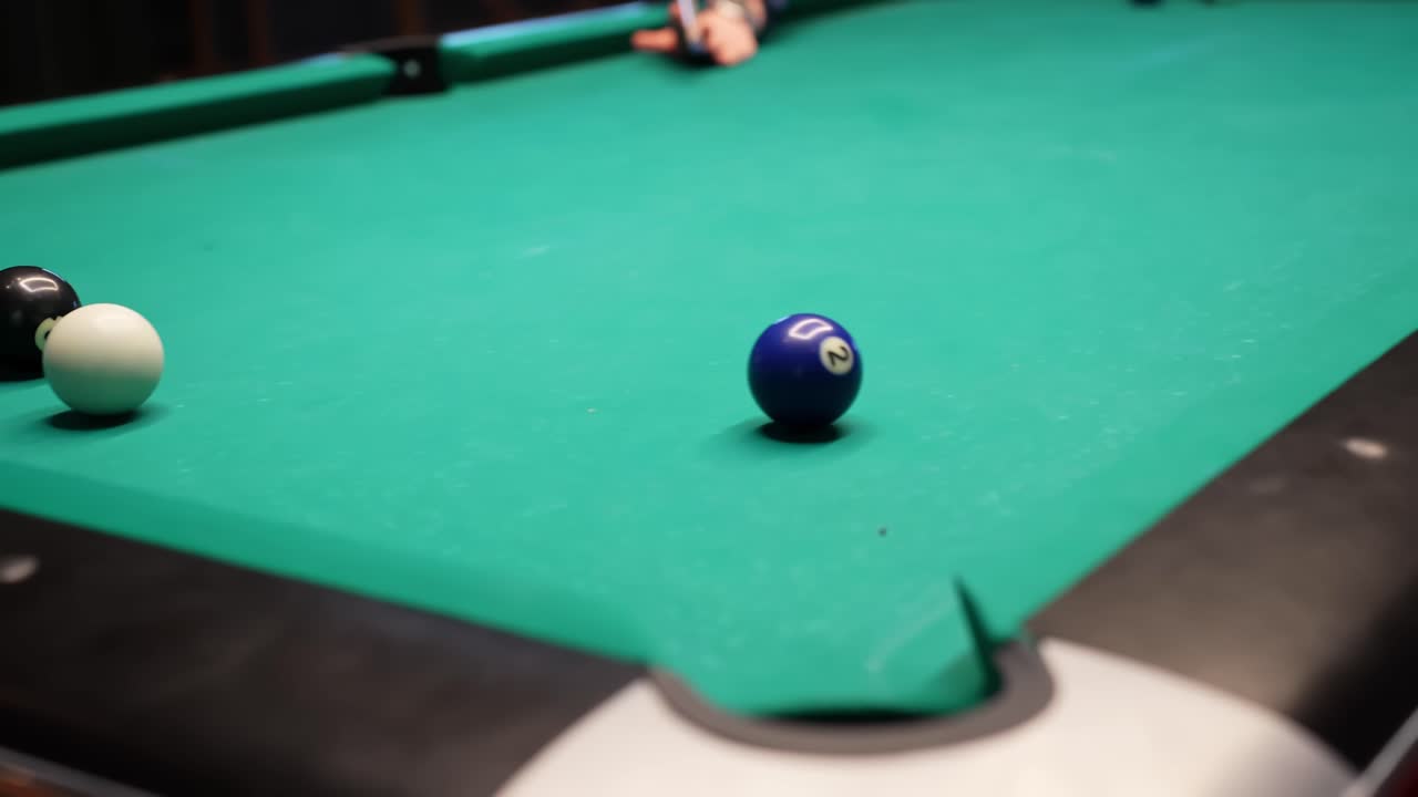Shallow-focus billiards scene as a player aims the cue ball toward a blue object near the side pocket on green felt, creating a tense, cinematic bar game moment