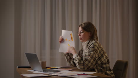 Trabajo-Remoto-Una-Mujer-Joven-Con-Gafas-Muestra-Un-Gráfico-En-Una-Cámara-Portátil-Mientras-Está-Sentada-En-Casa-En-La-Cocina-Realizando-Un-Informe-En-Línea.-Videollamada-Y-Conferencia-Con-Demostración-De-Horarios