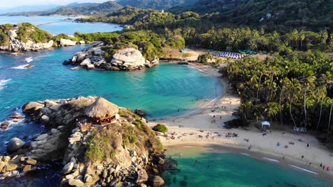 Aerial view overlooking shallow, blue water, rocks and people on the Playa Del Cabo beach on the coast of Tayrona national park, sunny evening, in Colombia - tilt up, drone shot