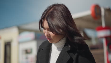 close-up of woman seated, focused on something, illuminated by sunlight, hair gently swaying in wind, looking away with a soft expression, background blurred with architectural features