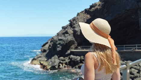 Una-Elegante-Mujer-Rubia-De-Verano-Con-Sombrero-Disfruta-Del-Clima-Atlántico,-Vista-Posterior