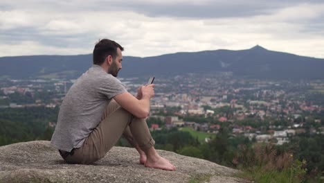 hombre caucásico usando un teléfono móvil afuera, escribiendo y deslizando el dedo, sentado en una roca con una vista impresionante en segundo plano