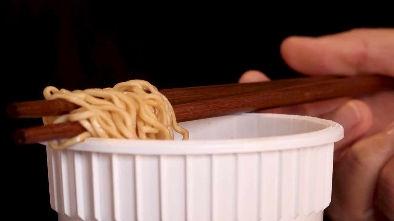 A hand skillfully lifts and lowers cooked instant noodles with wooden chopsticks over a white cup, set against a dark background with soft, even lighting