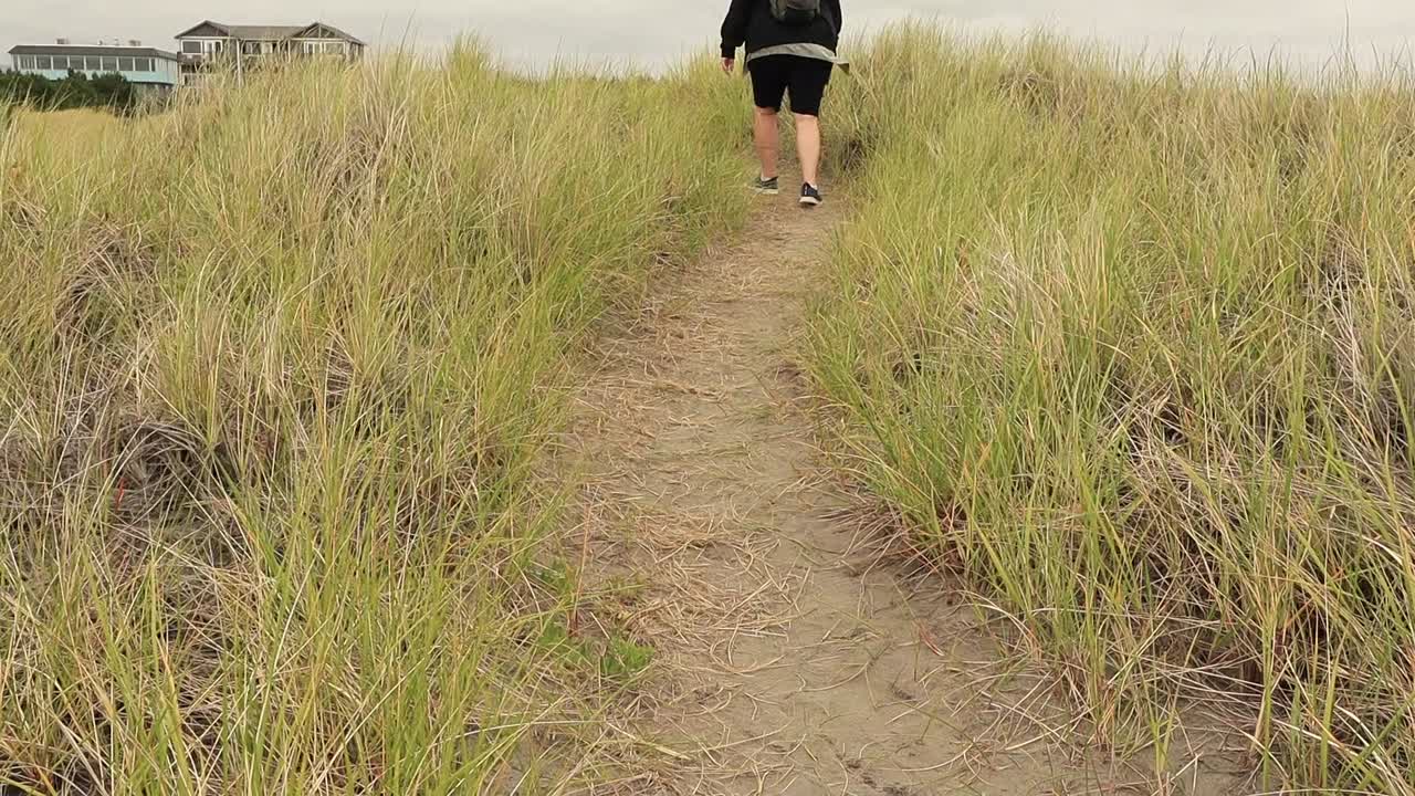Dunes path woman walking only showing feet in sneakers up to waist shot pan up to show coastal buildings in the background