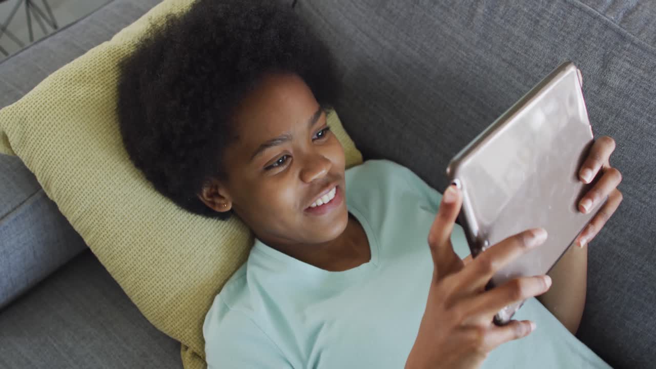 Happy african american teenage girl lying on sofa, using tablet