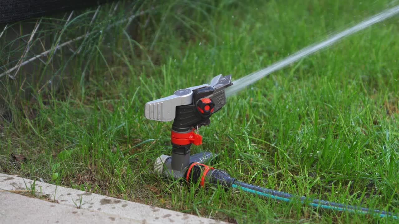 Orange gray impulse sprinkler in a lawn corner with green garden tube waters the lawn, view from diagonal above near ground level