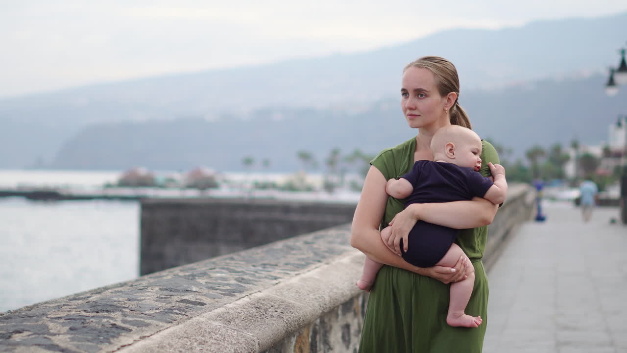 On a historic European square near the ocean, a young mother walks with her baby son, appreciating the waves' sight and sharing joyful smiles