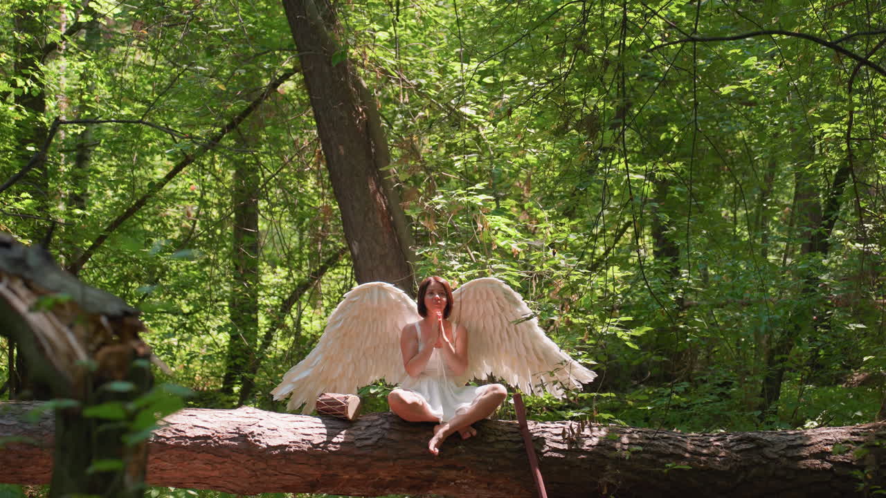 Mythical being with angelic wings in white dress kneels on fallen forest log praying to gods under soft sunlight, surrounded by green foliage, expressing reverence