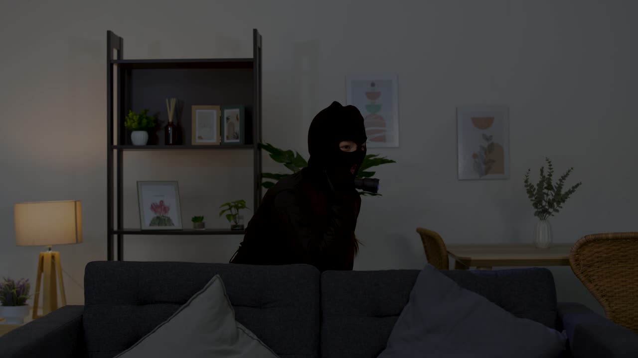 A masked intruder cautiously moves through a dark living room using a flashlight at night
